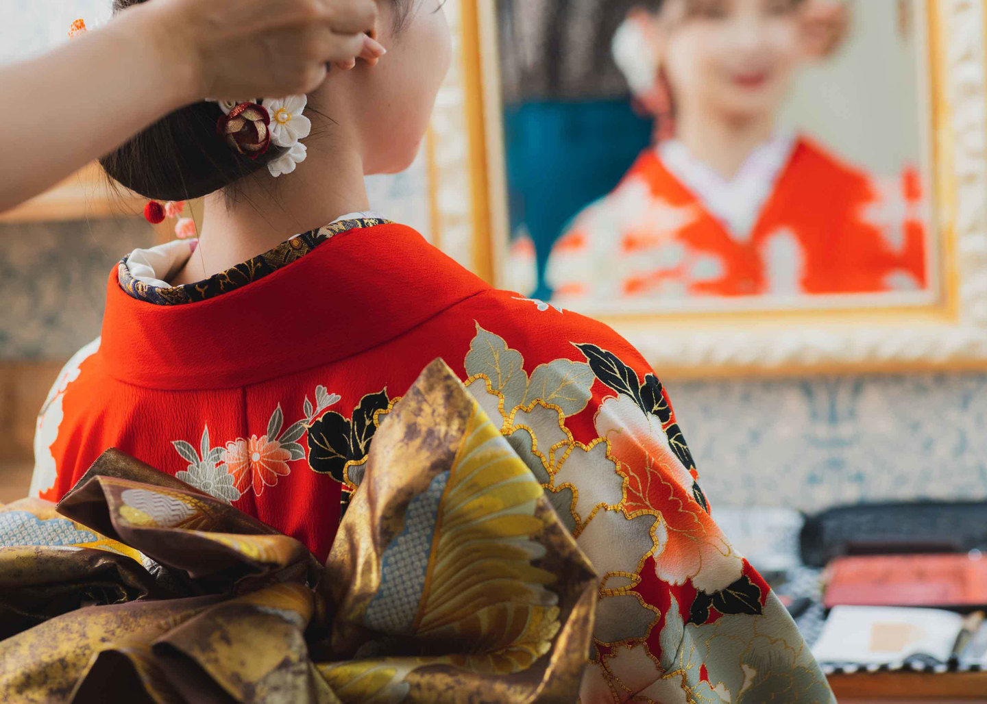 三原市・冨士写真館での成人式当日のヘアセット。鏡越しに笑顔を見せる赤い振袖姿のお嬢様と、プロの手による丁寧なヘアセット・髪飾りの取り付け風景。